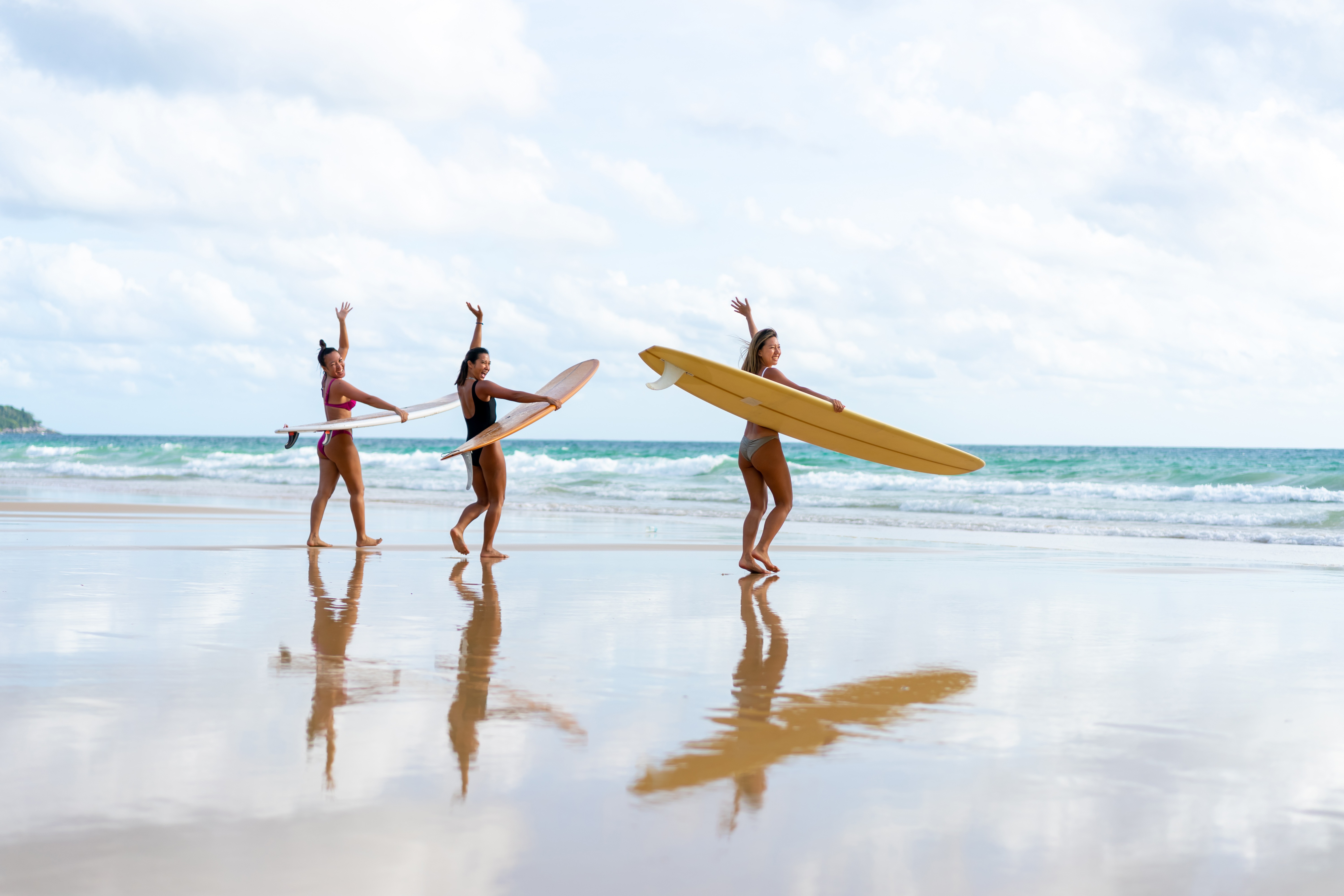 3 Frauen mit Surfboards winken in die Kamera und gehen Richtung Meer