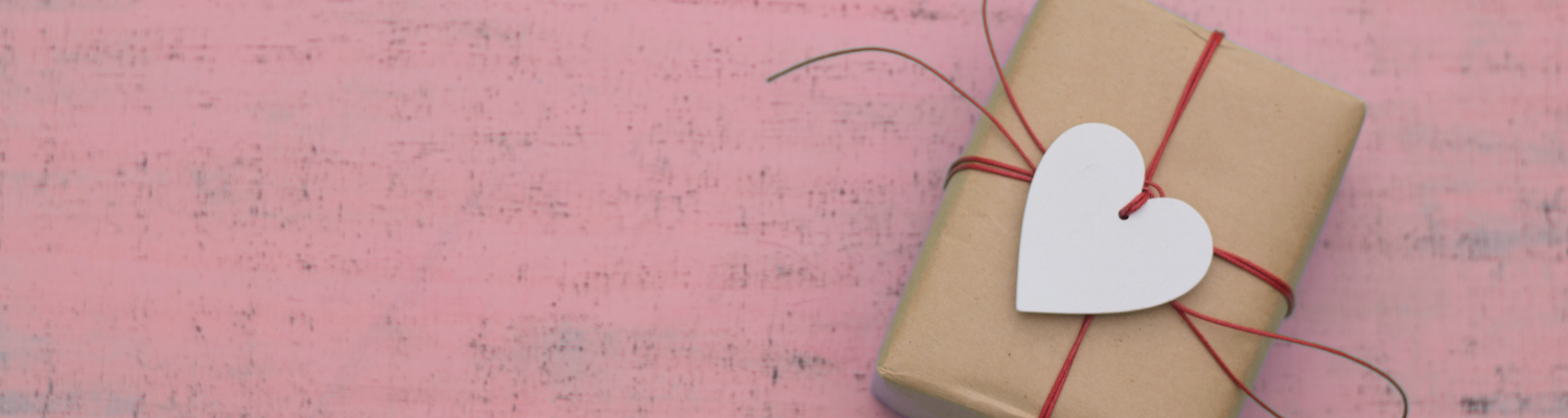 Natürlich verpacktes Geschenk mit rotem Band und Holzherz auf rosa Hintergrund – symbolisches Bild für persönliche, herzliche Weihnachtsgeschenke für Hundehalter.